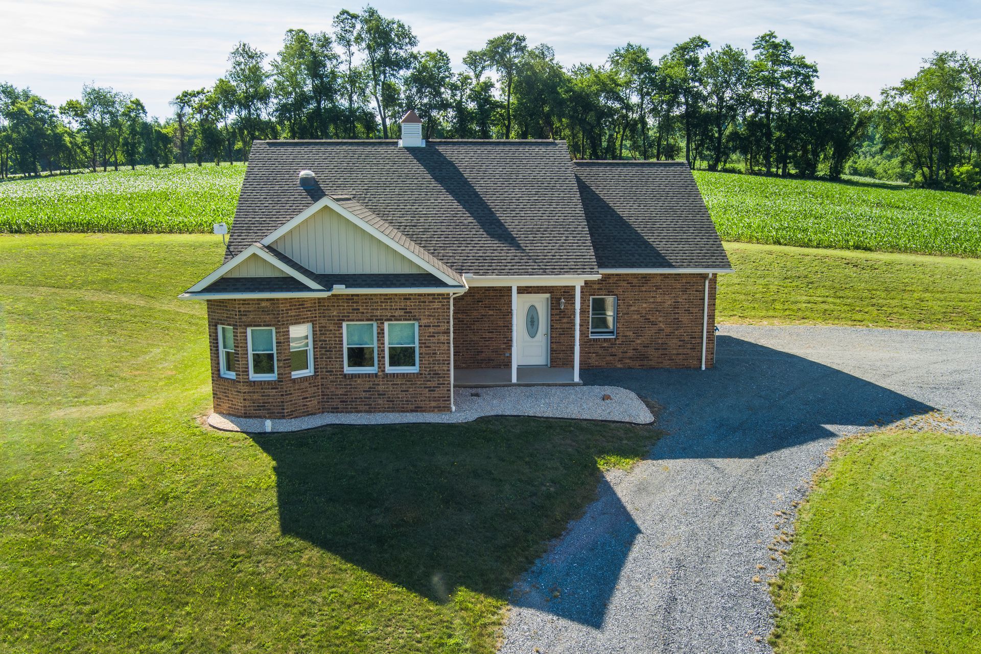 Small brick house with gray roof and gravel driveway in a green rural field