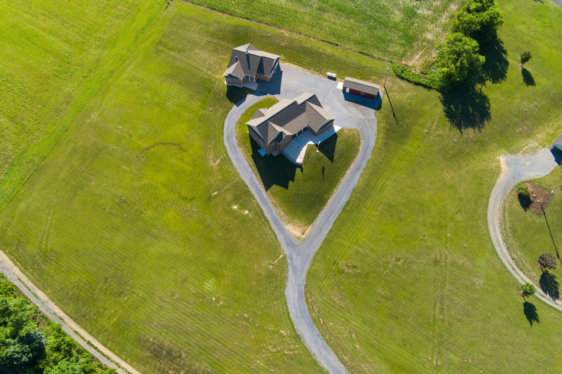 Aerial view of a house and driveway on a large grassy property, with a small outbuilding nearby.