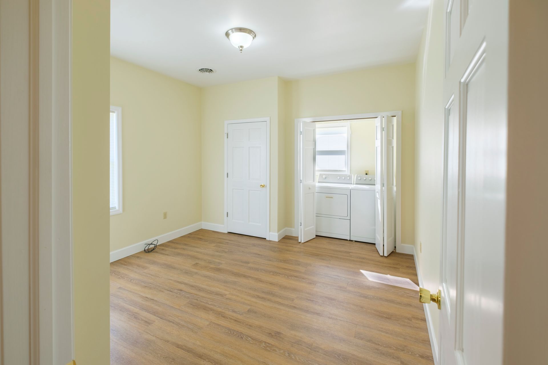 Empty beige bedroom with hardwood floors, white closet doors, and a window with curtains