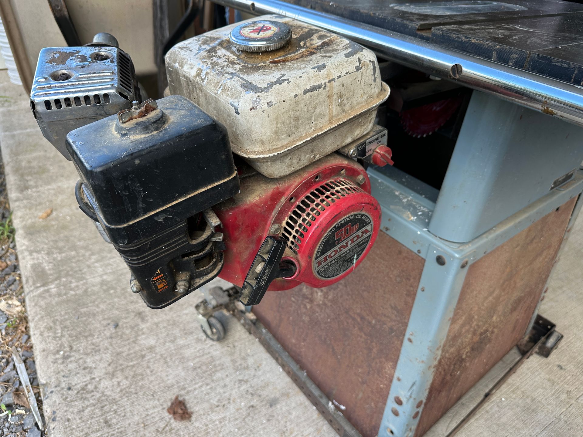Old gasoline engine mounted on a metal cart, with red recoil starter and black and white fuel tanks.