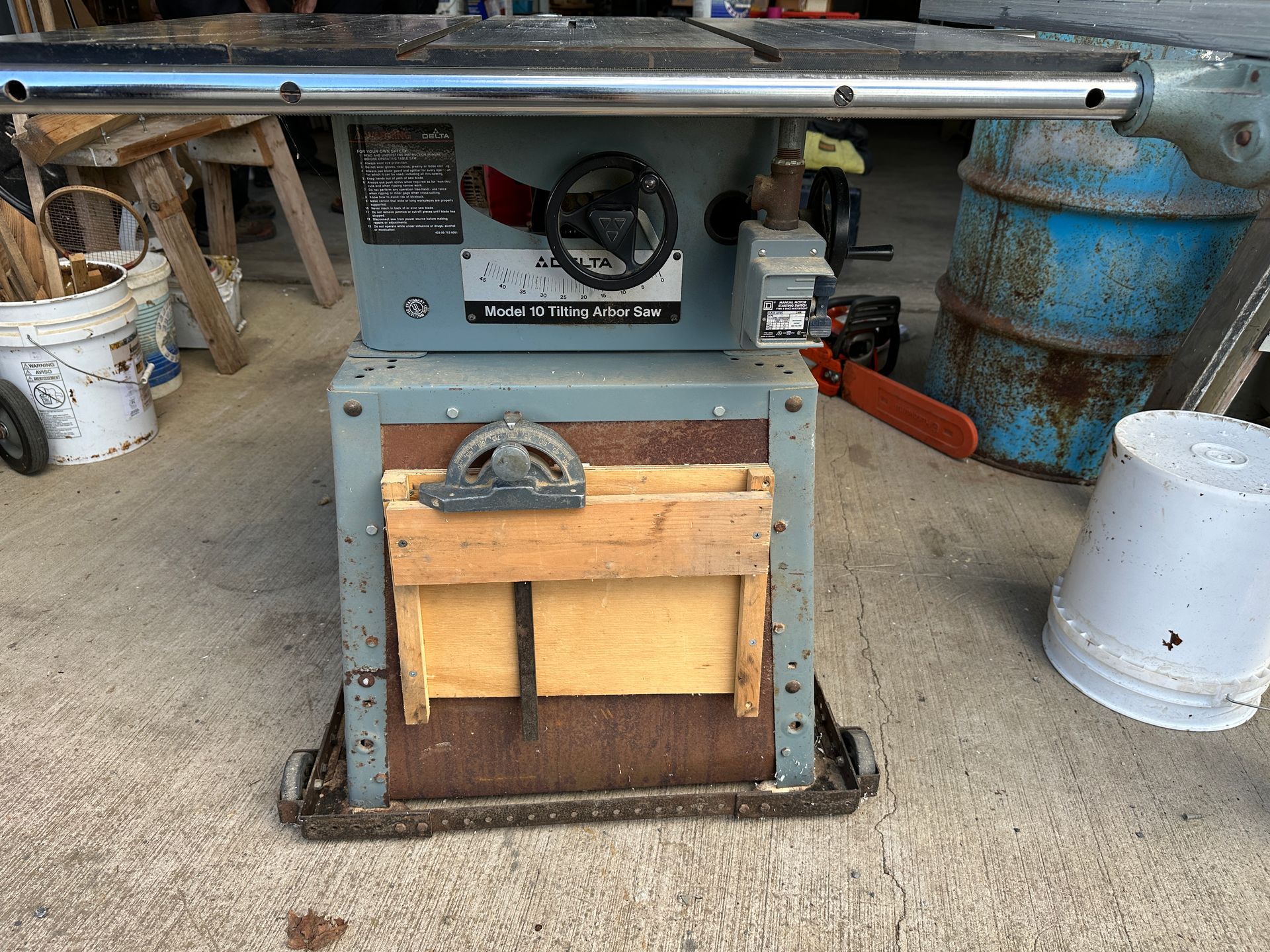 Workshop machine with blue frame and wooden base, sitting on a gravel floor with tools and barrels nearby