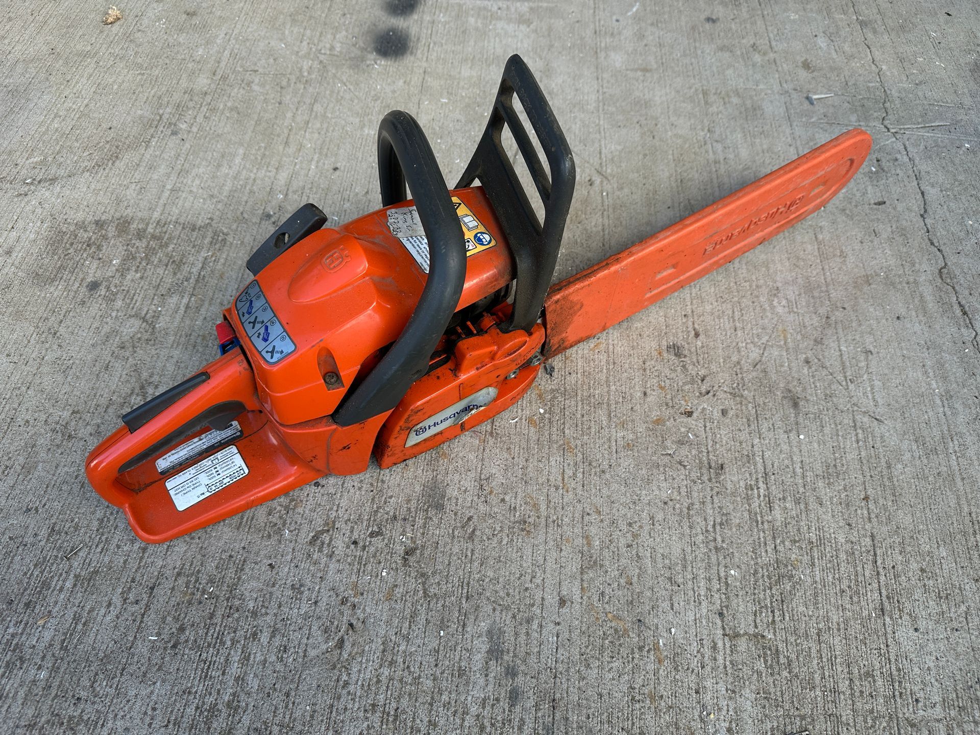 Orange chainsaw lying on concrete, with black handle and long orange guide bar.