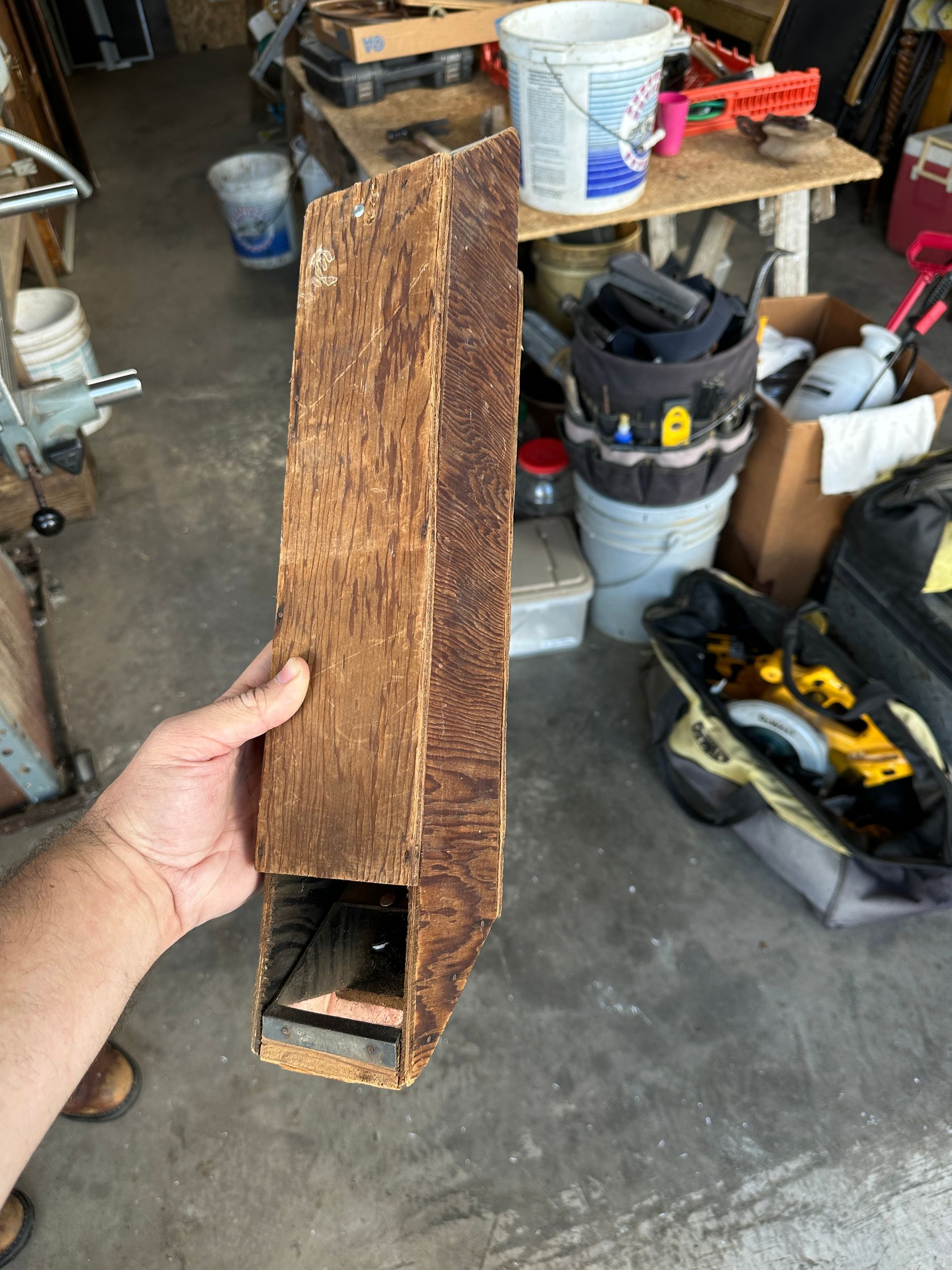 Hand holding a rough wooden beam with a rectangular cutout, in a cluttered garage.