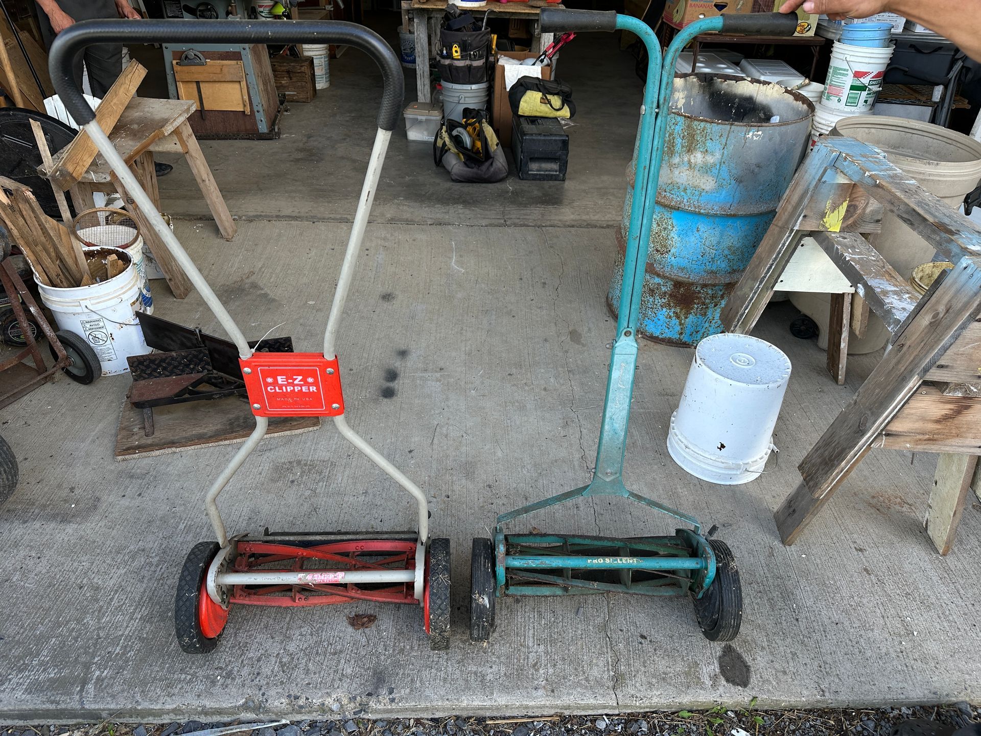 Two push lawn mowers and yard tools in a garage or workshop, with a blue gas cylinder nearby