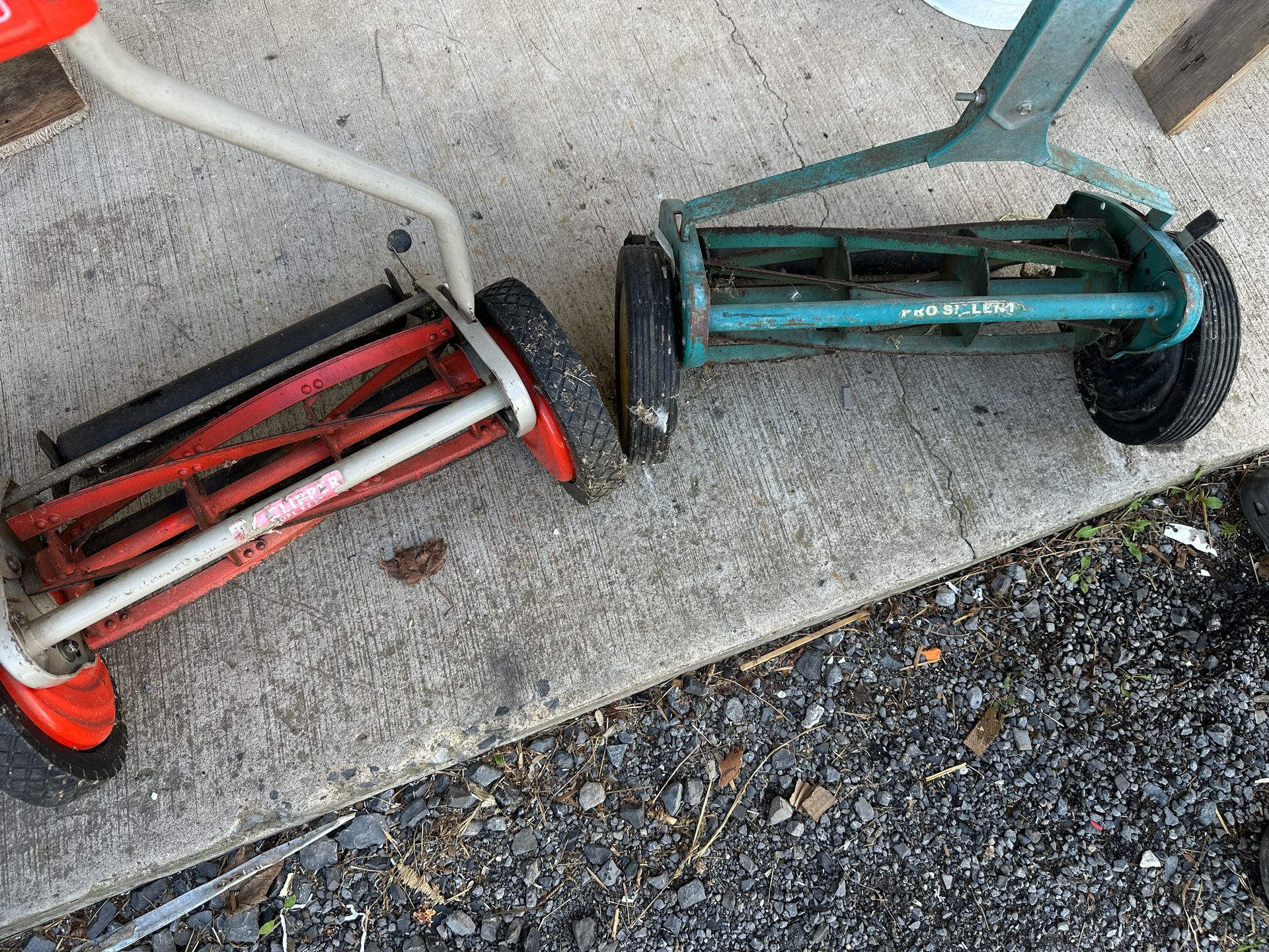 Two old lawn mowers, red and teal, lying on a concrete curb beside gravel.