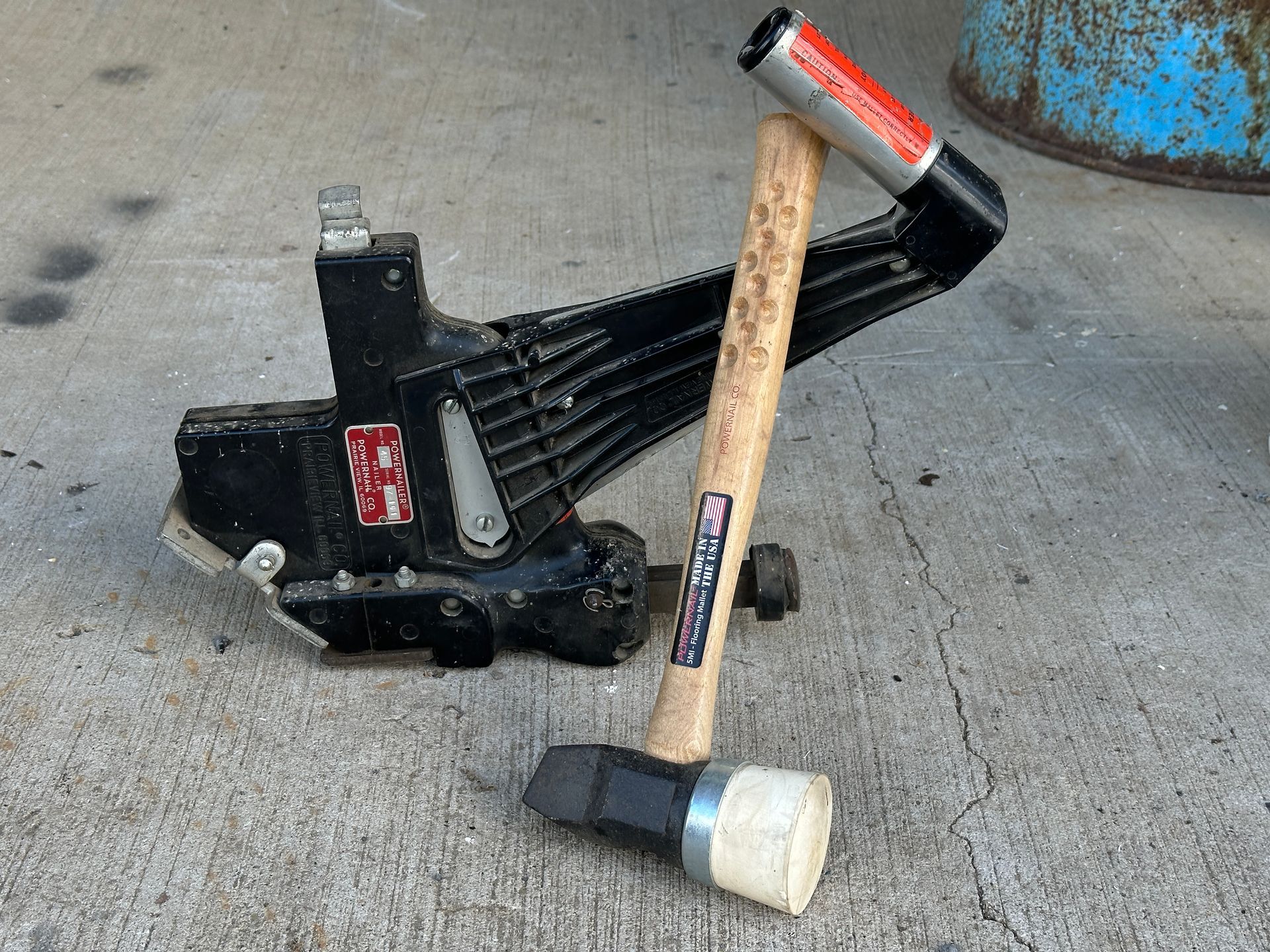 Black floor jack and hammer on concrete beside a blue barrel.