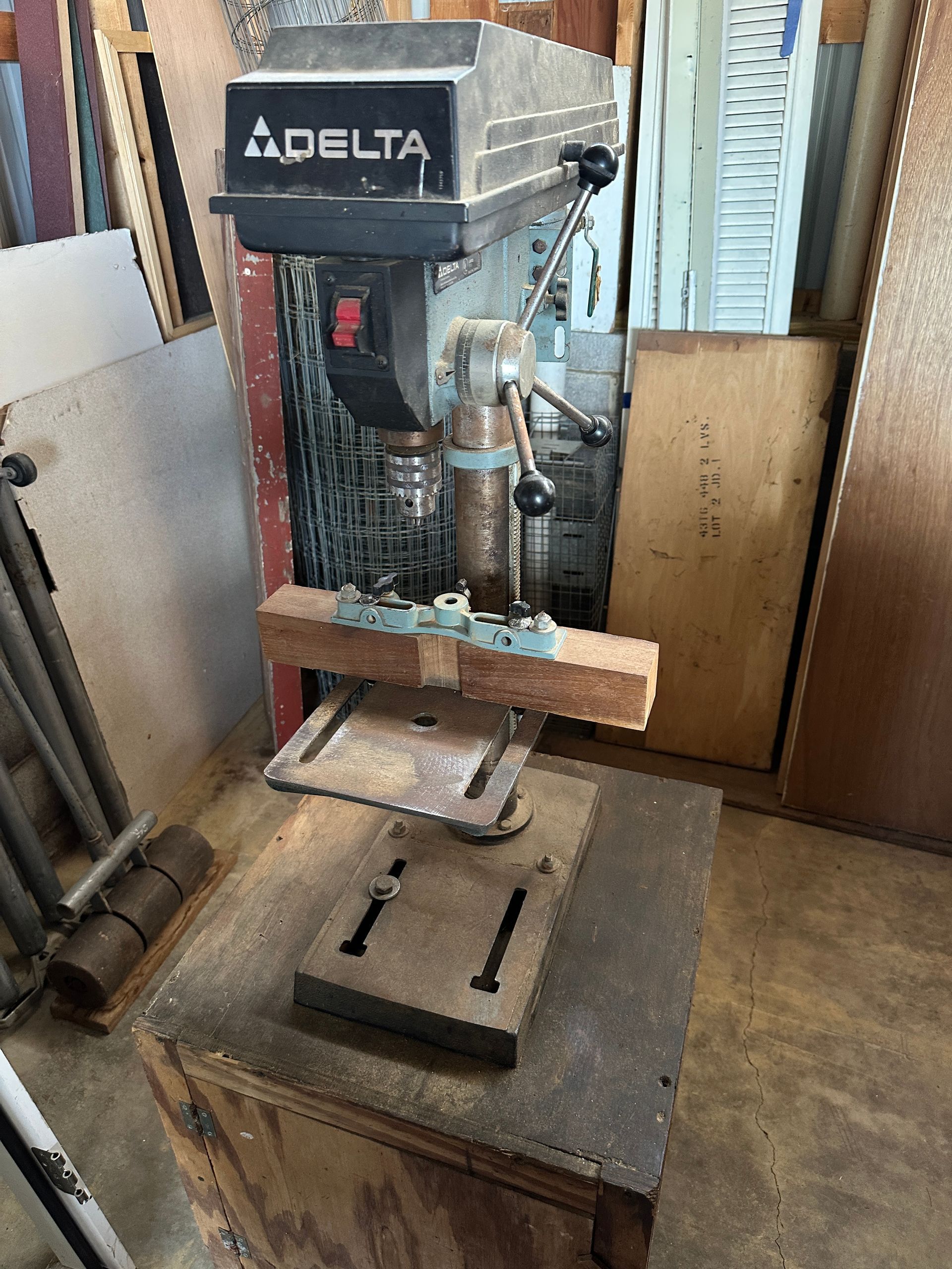Delta drill press in a workshop, with a wooden board clamped on the table and tools below.