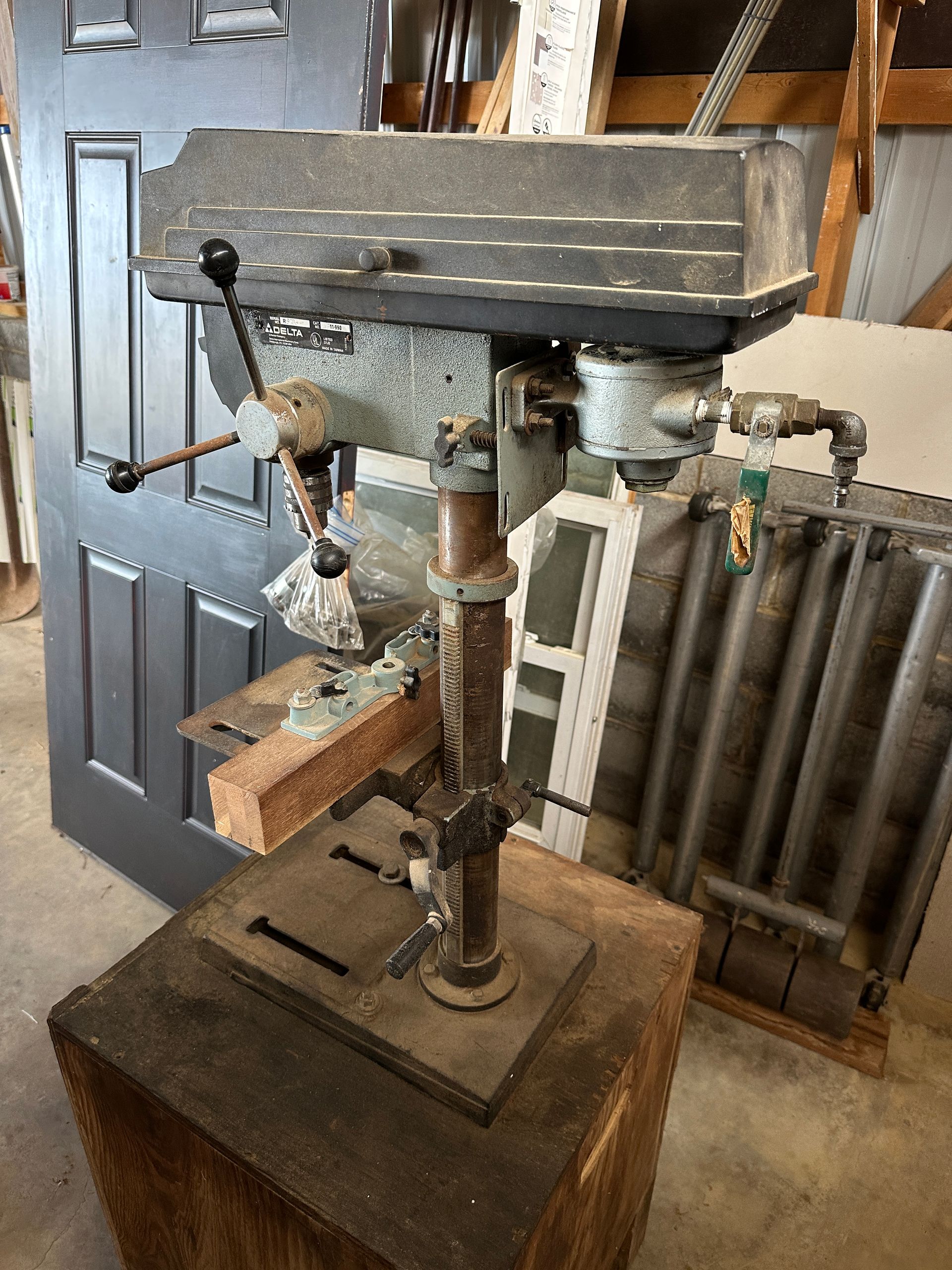 Old, dust-covered drill press in a workshop beside a door and corrugated metal wall