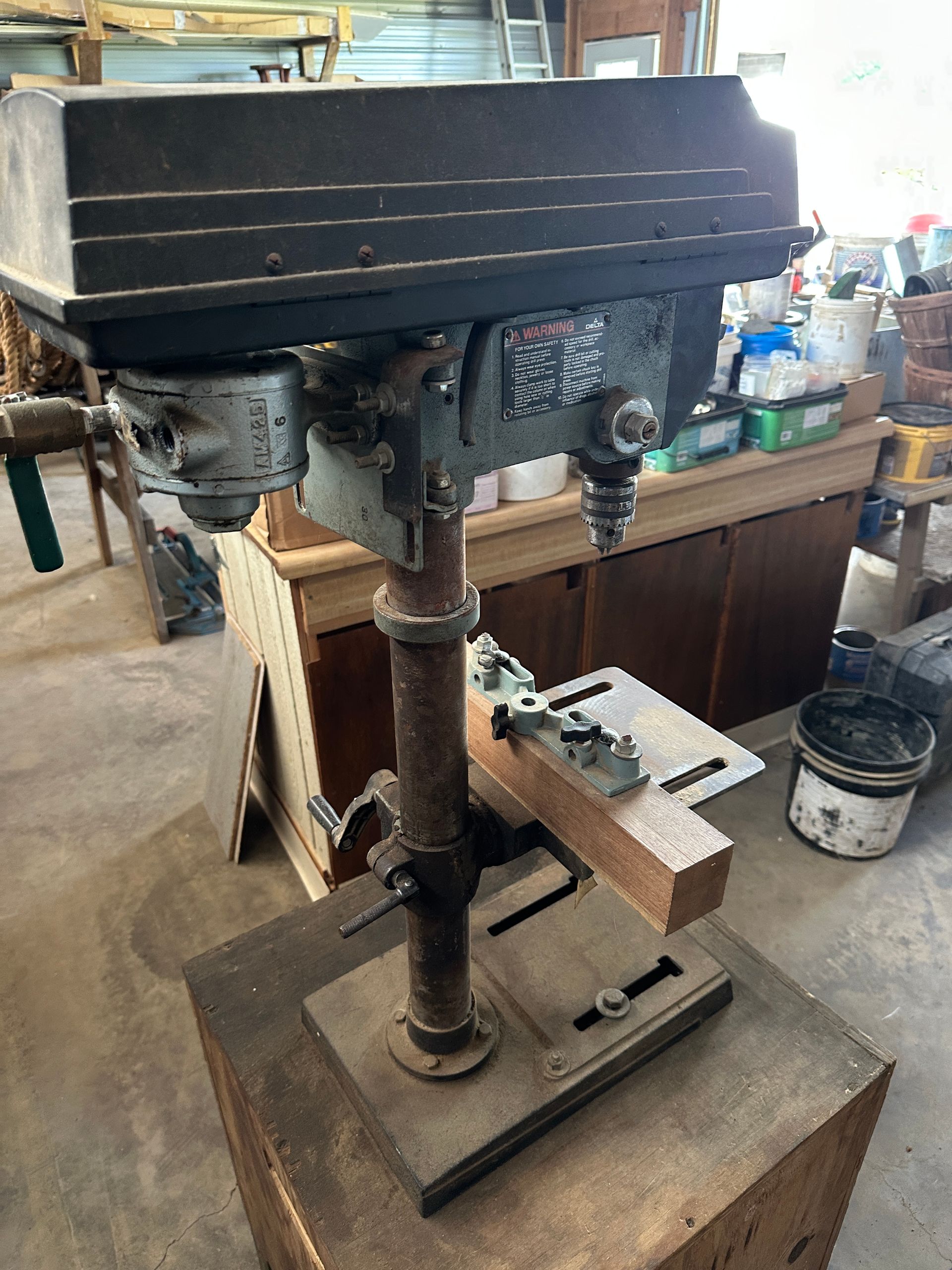 Old floor drill press in a workshop with a black head, metal column, and wooden base.