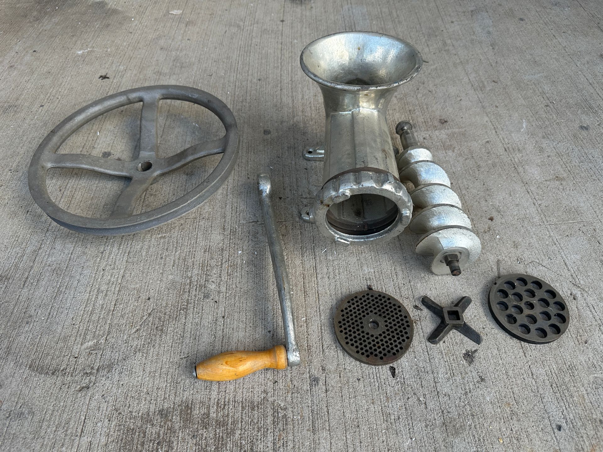 Metal meat grinder with hand crank, attachments, and plates laid out on a concrete surface.