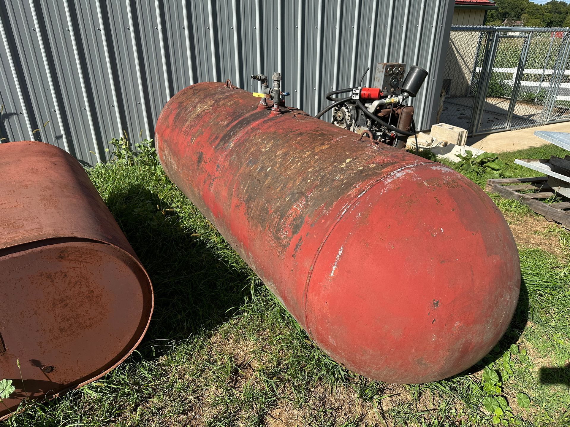 Rusty red metal tank lying on grass beside a corrugated fence.