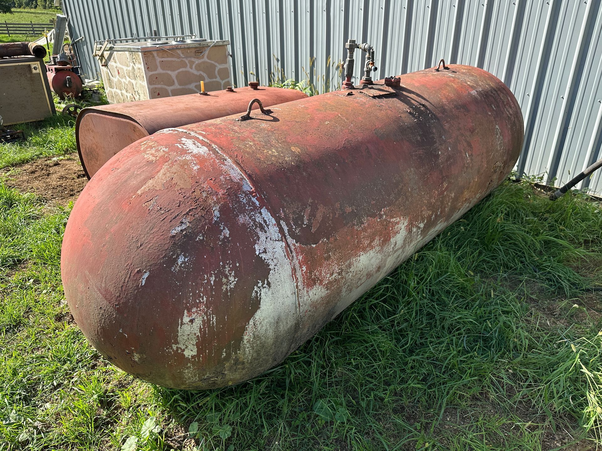 Rusty red horizontal storage tank in a grassy yard beside a corrugated metal fence