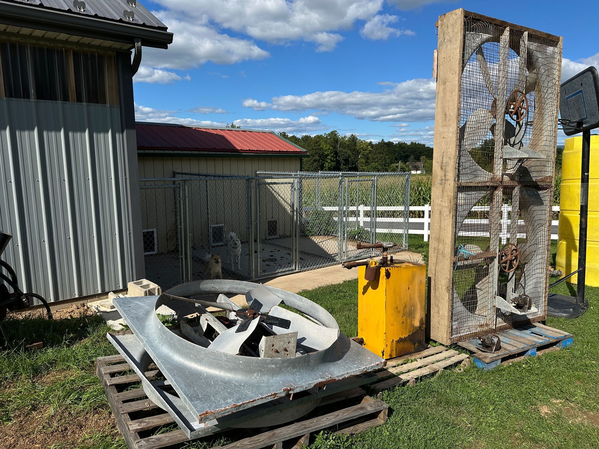 Outdoor yard with a large metal fan, yellow equipment, and a rusted mesh structure beside a building
