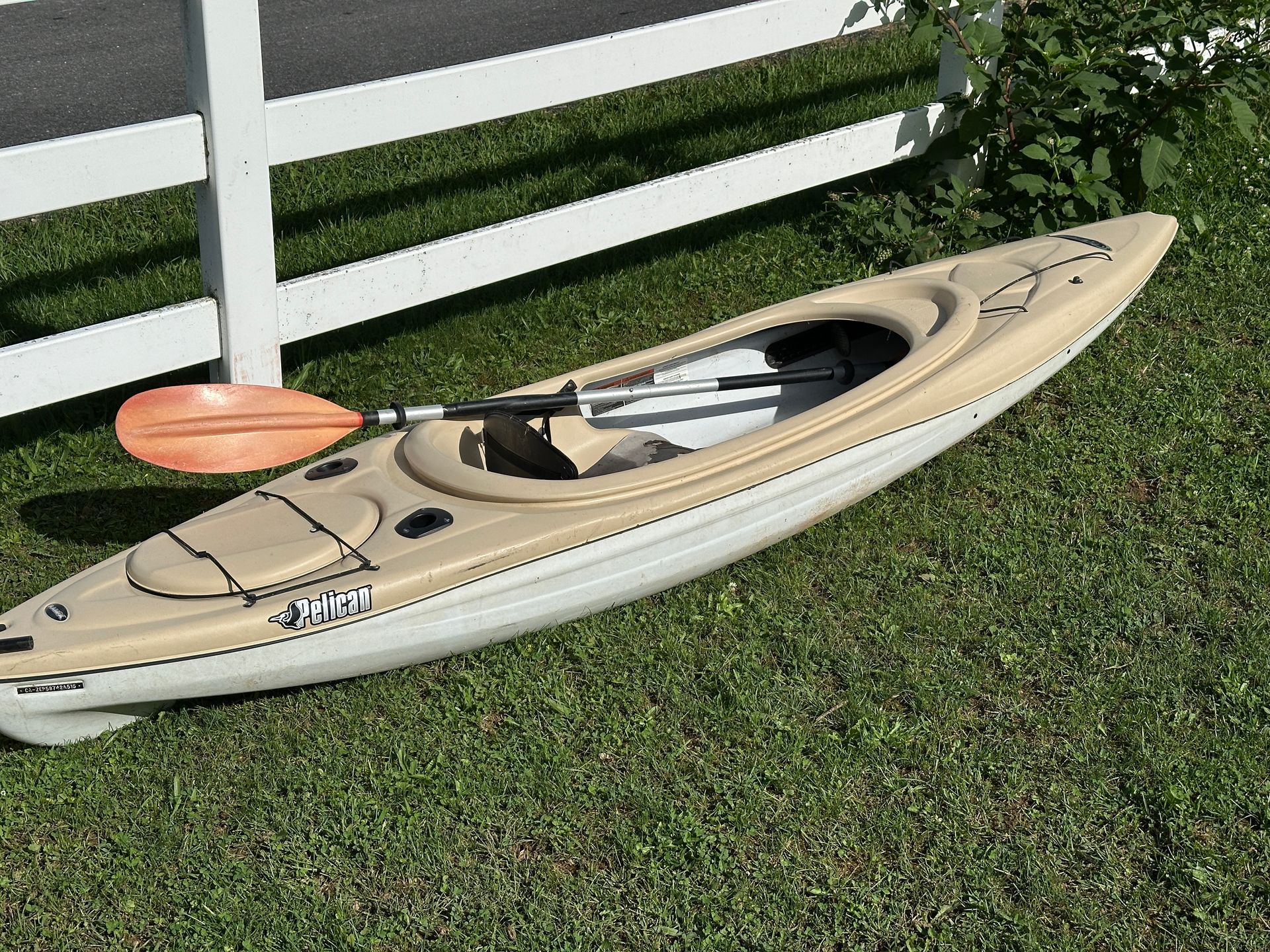 Tan kayak with orange paddle resting on grass beside a white fence