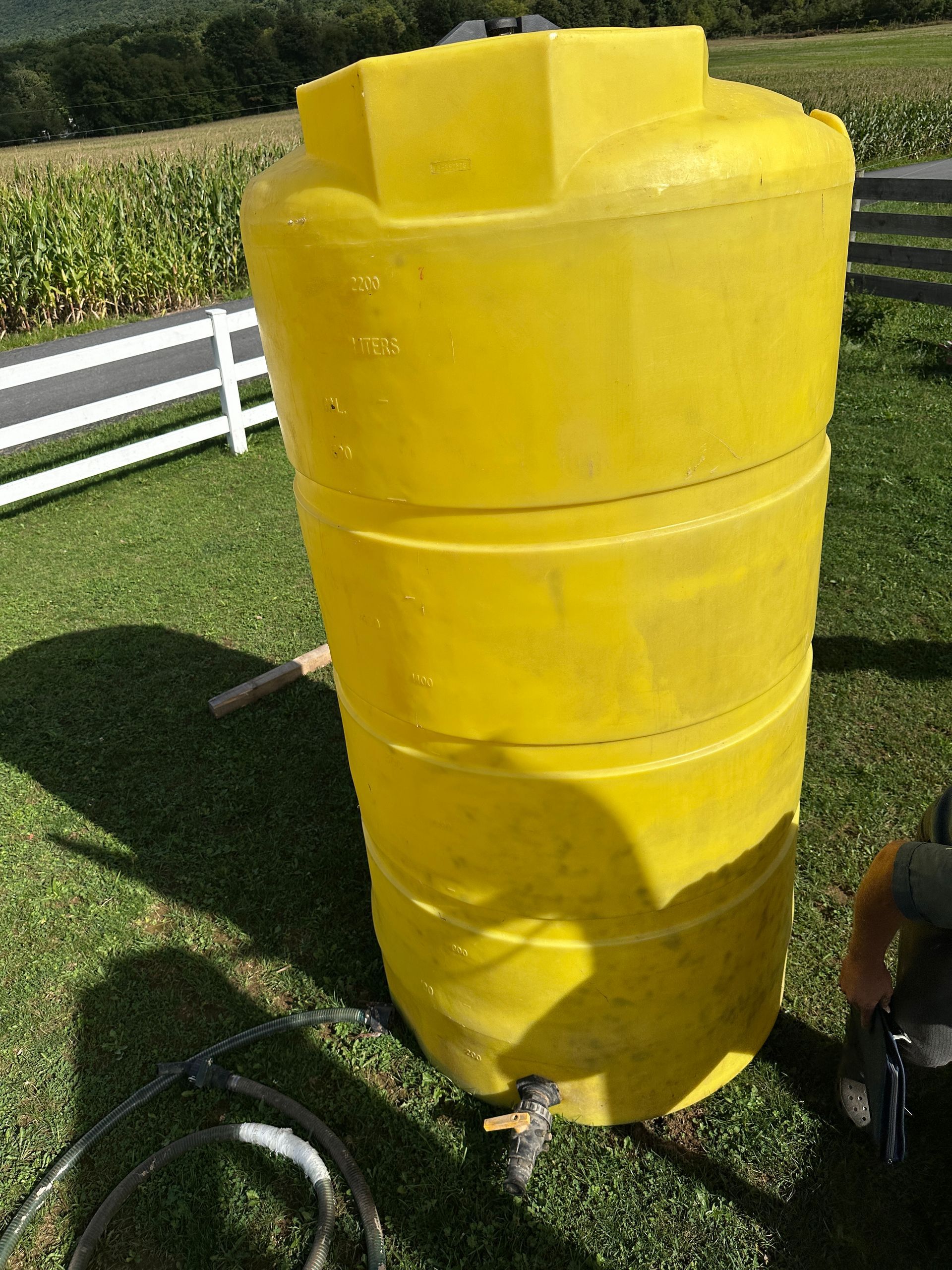 Yellow cylindrical tank or barrel standing outdoors on grass near a white fence