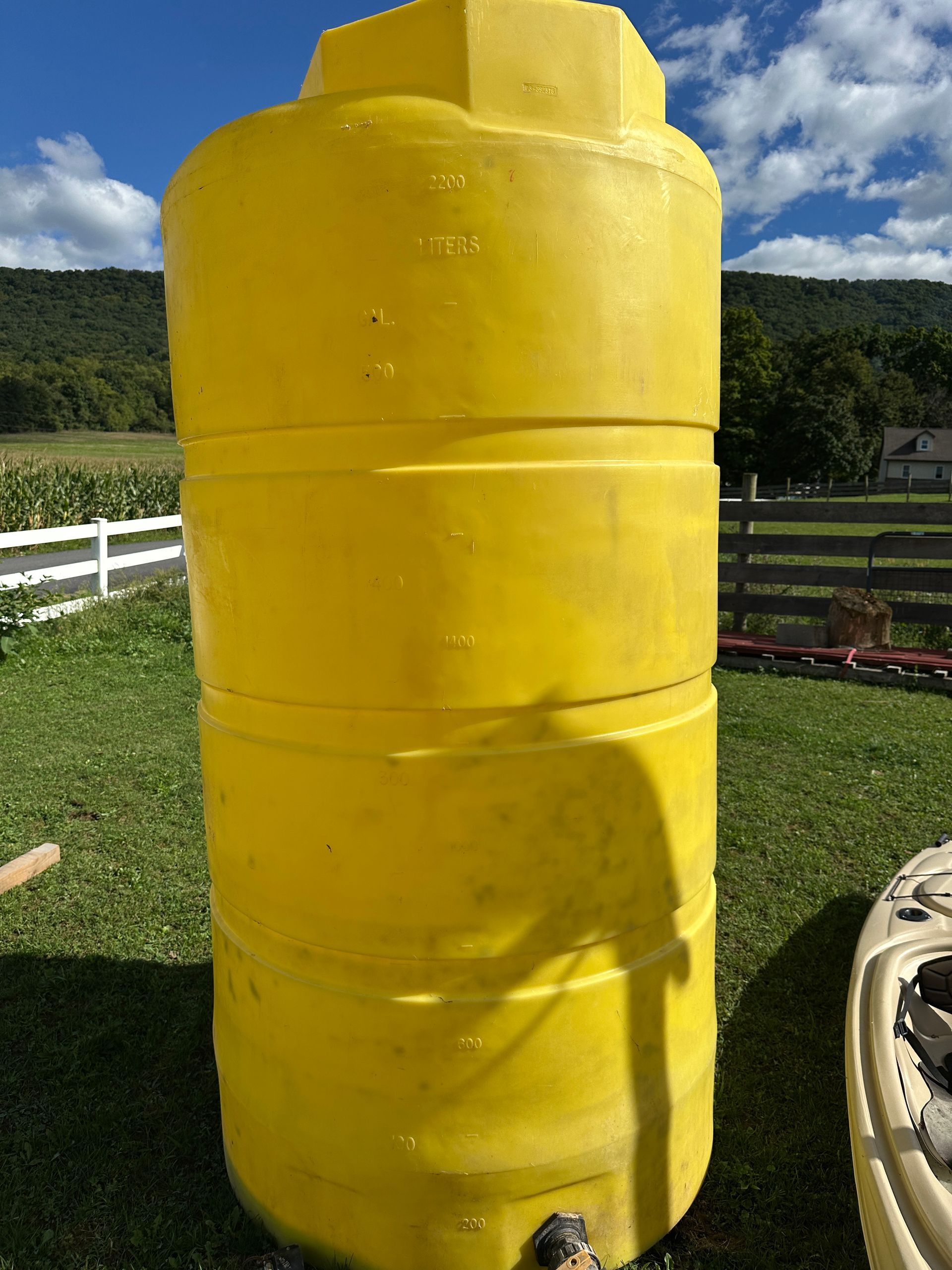 Yellow plastic tank standing outdoors on grass with white fence and hills in the background
