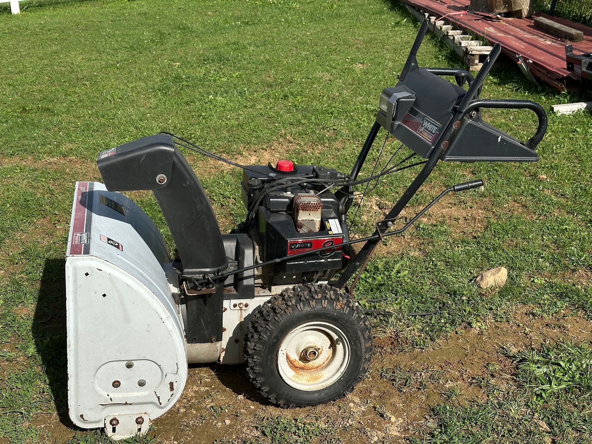 Snow blower with black handle and gray chute parked on grass near a wooden deck.