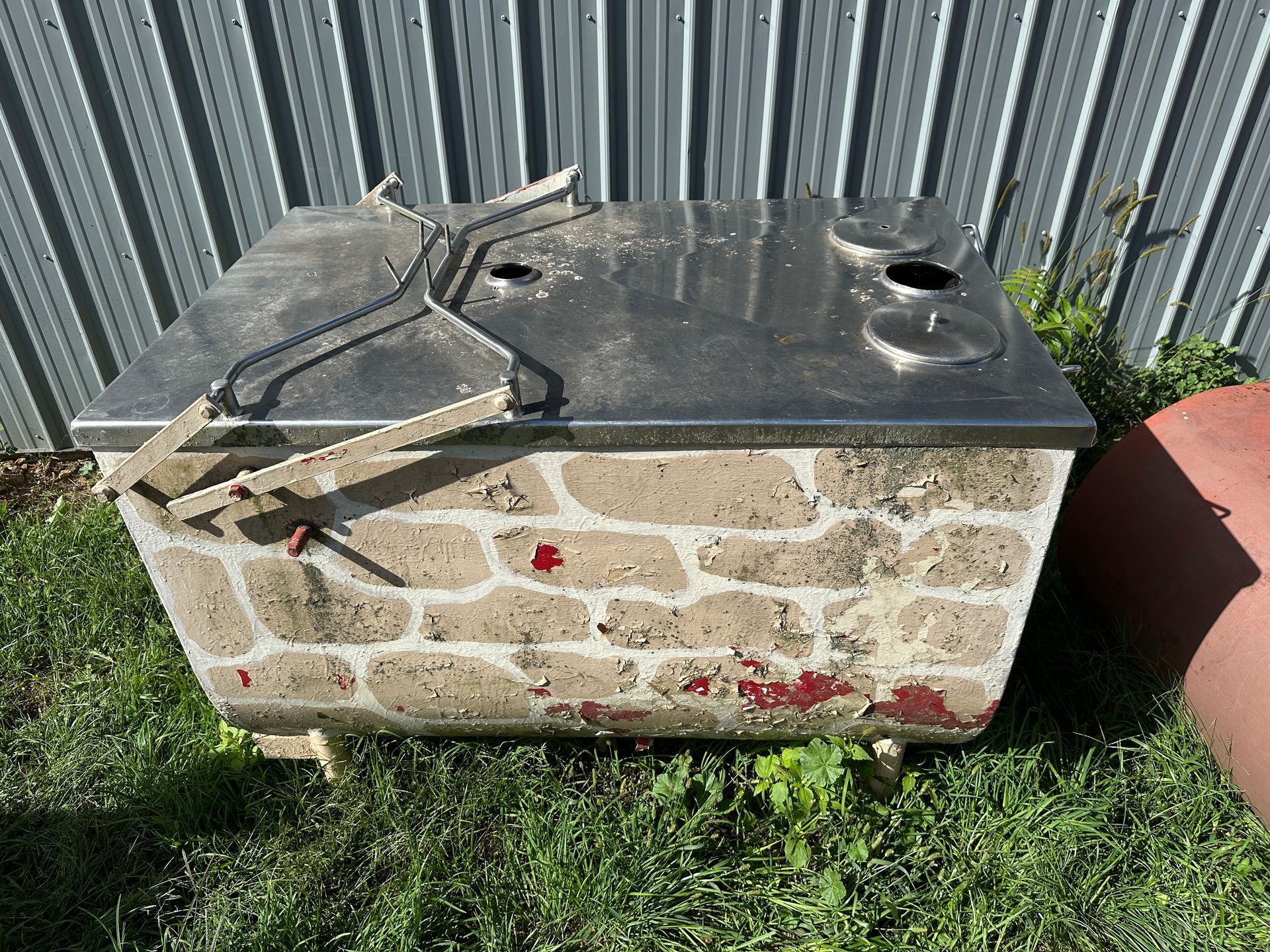 Stone utility box with black lid and hose, sitting on grass by a metal fence and red planter