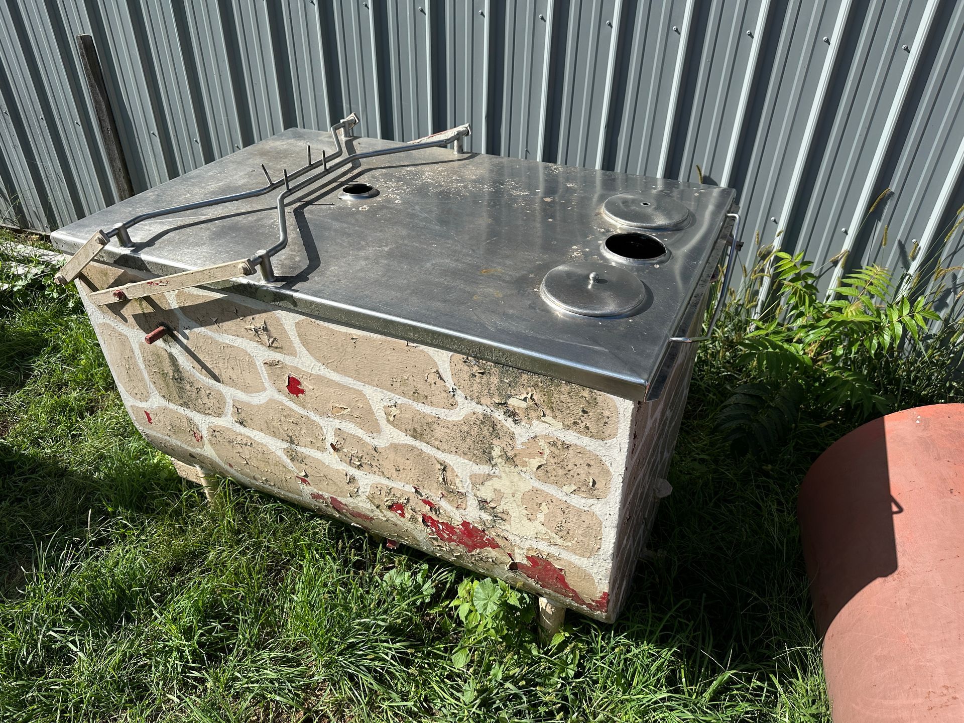 Stone utility box with black metal top, cables, and red stains beside a corrugated fence and grass.