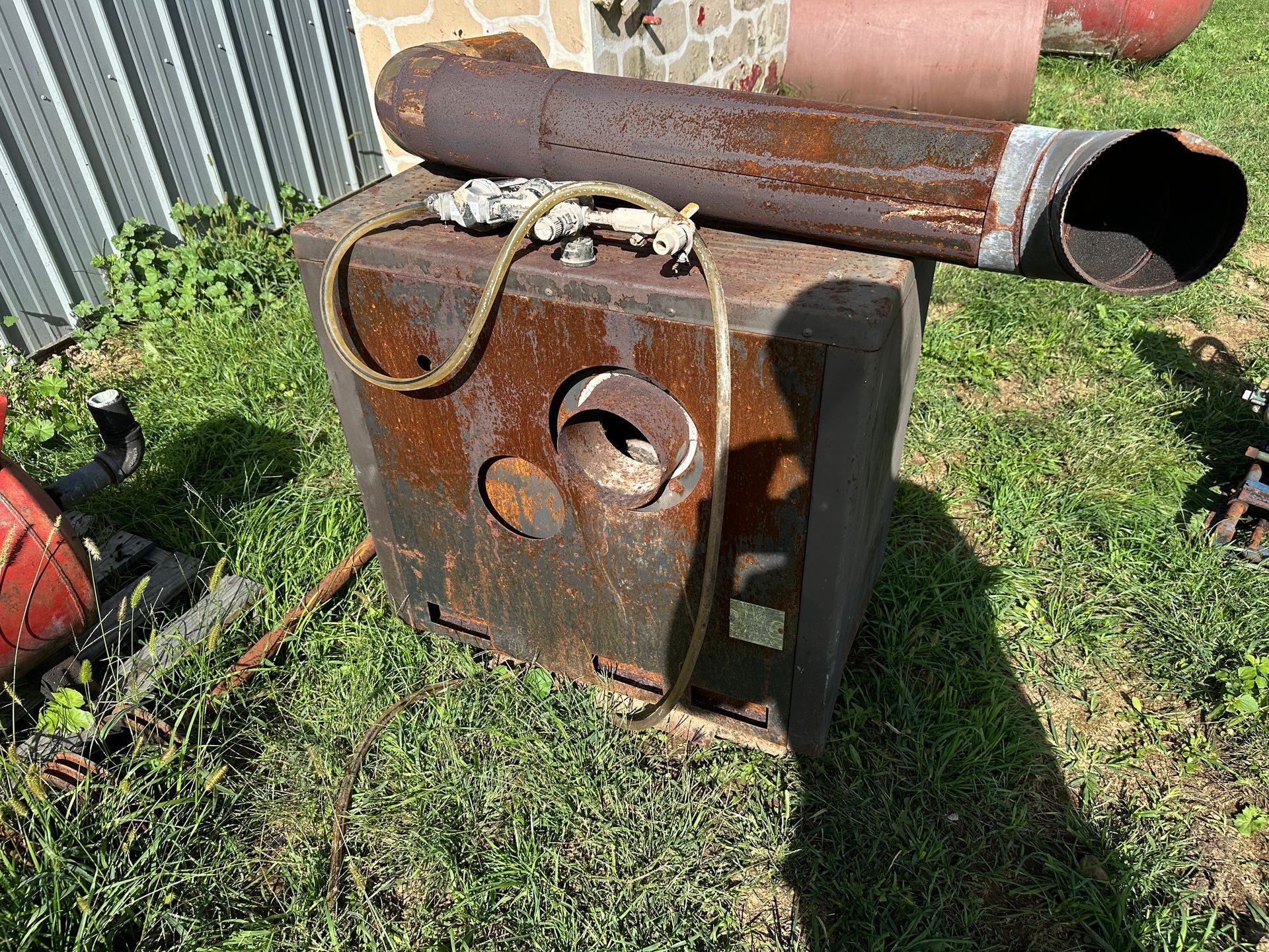 Rusty metal box with a circular vent and pipe attached, sitting on grass outdoors.