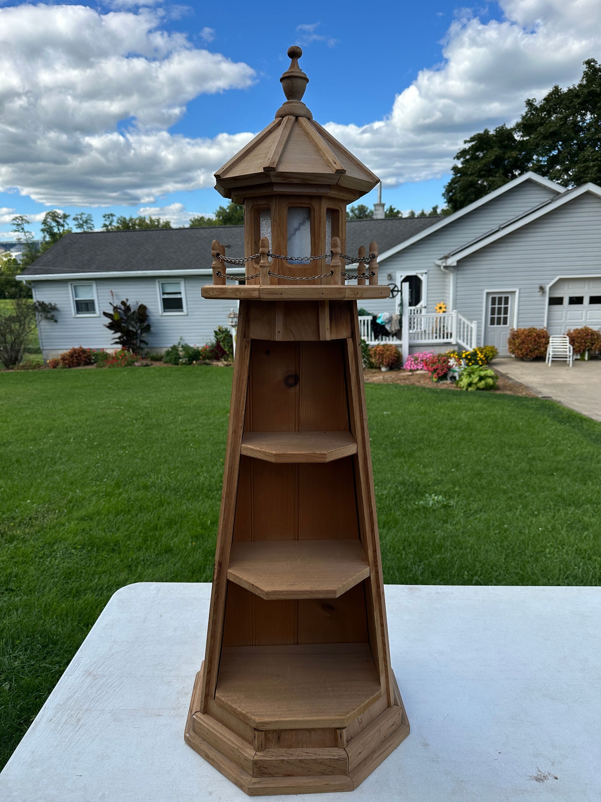 Tall wooden outdoor shelf or birdhouse on a white platform in a grassy yard, with houses and flowers behind.