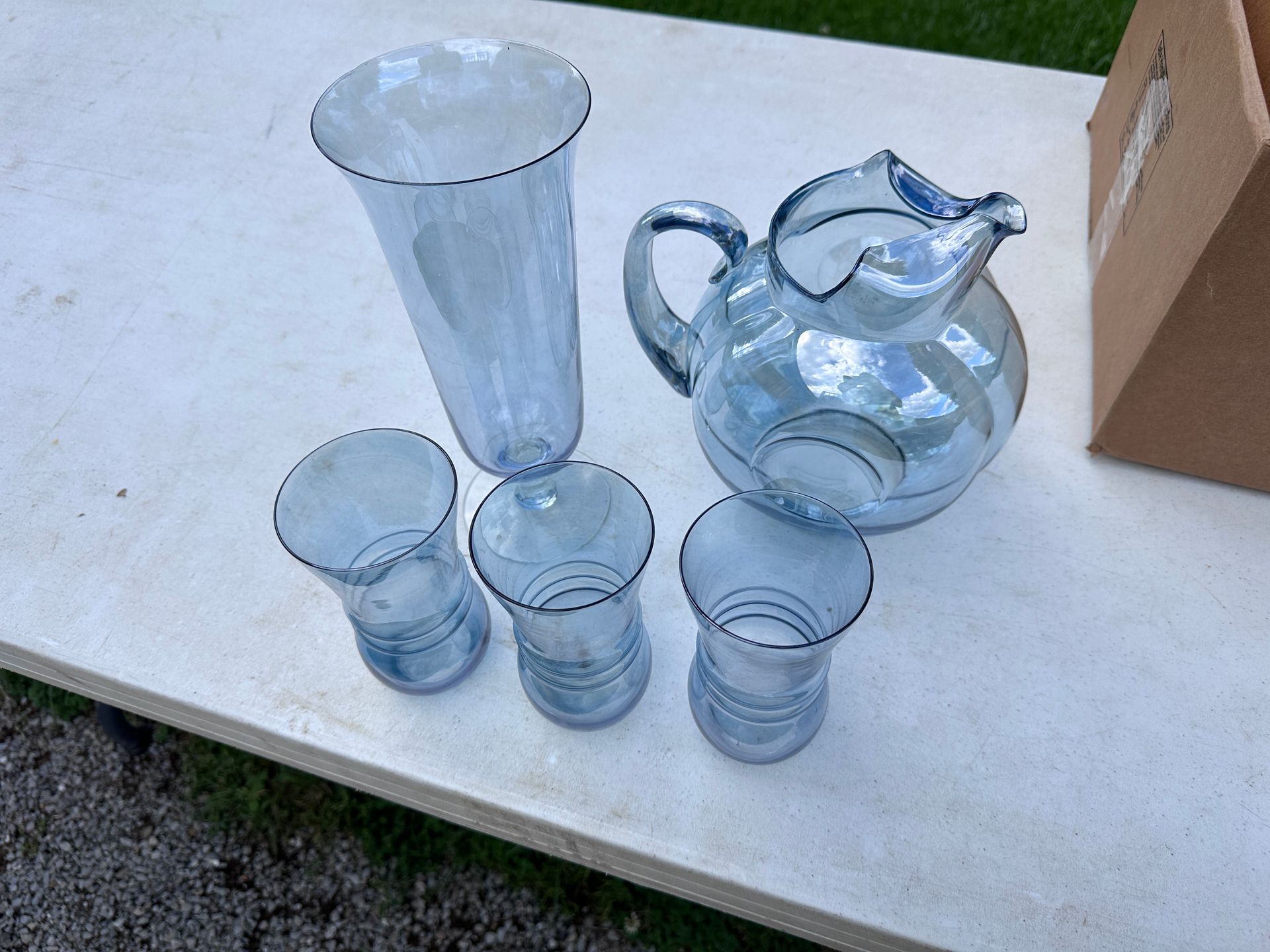 Clear glass pitcher, tall vase, and three glasses on a white table outdoors.