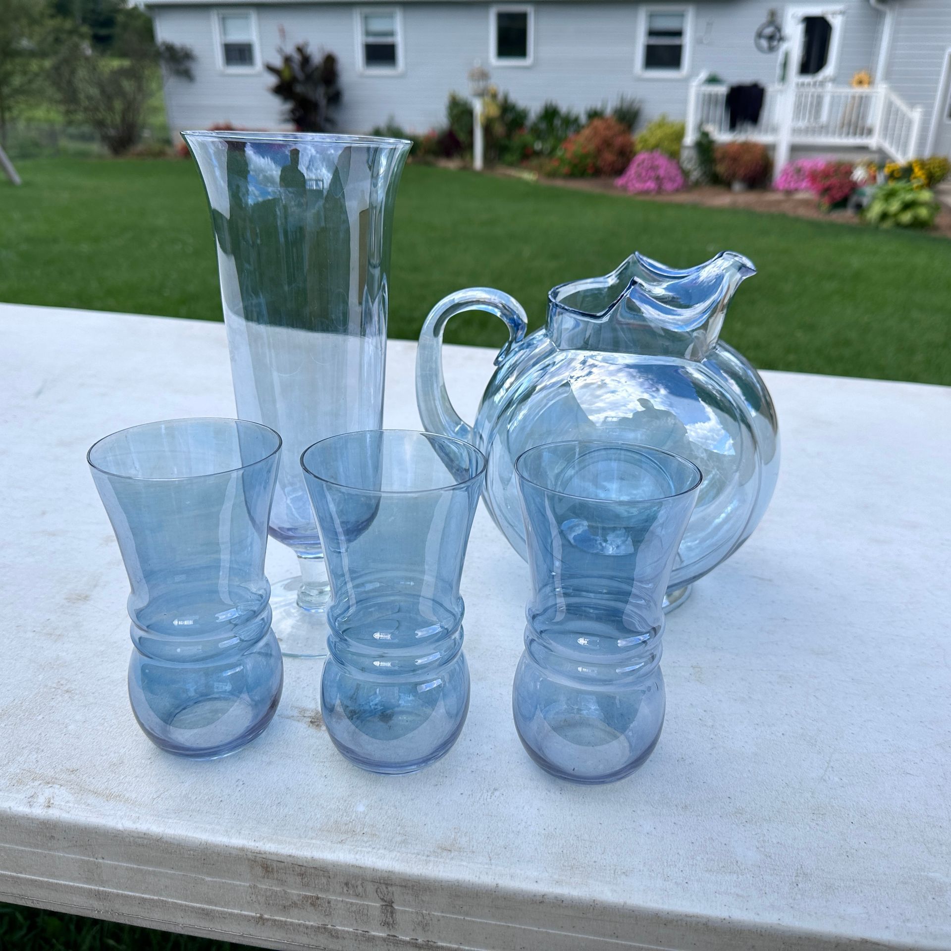Four translucent blue glass vases and cups arranged on a white table outdoors