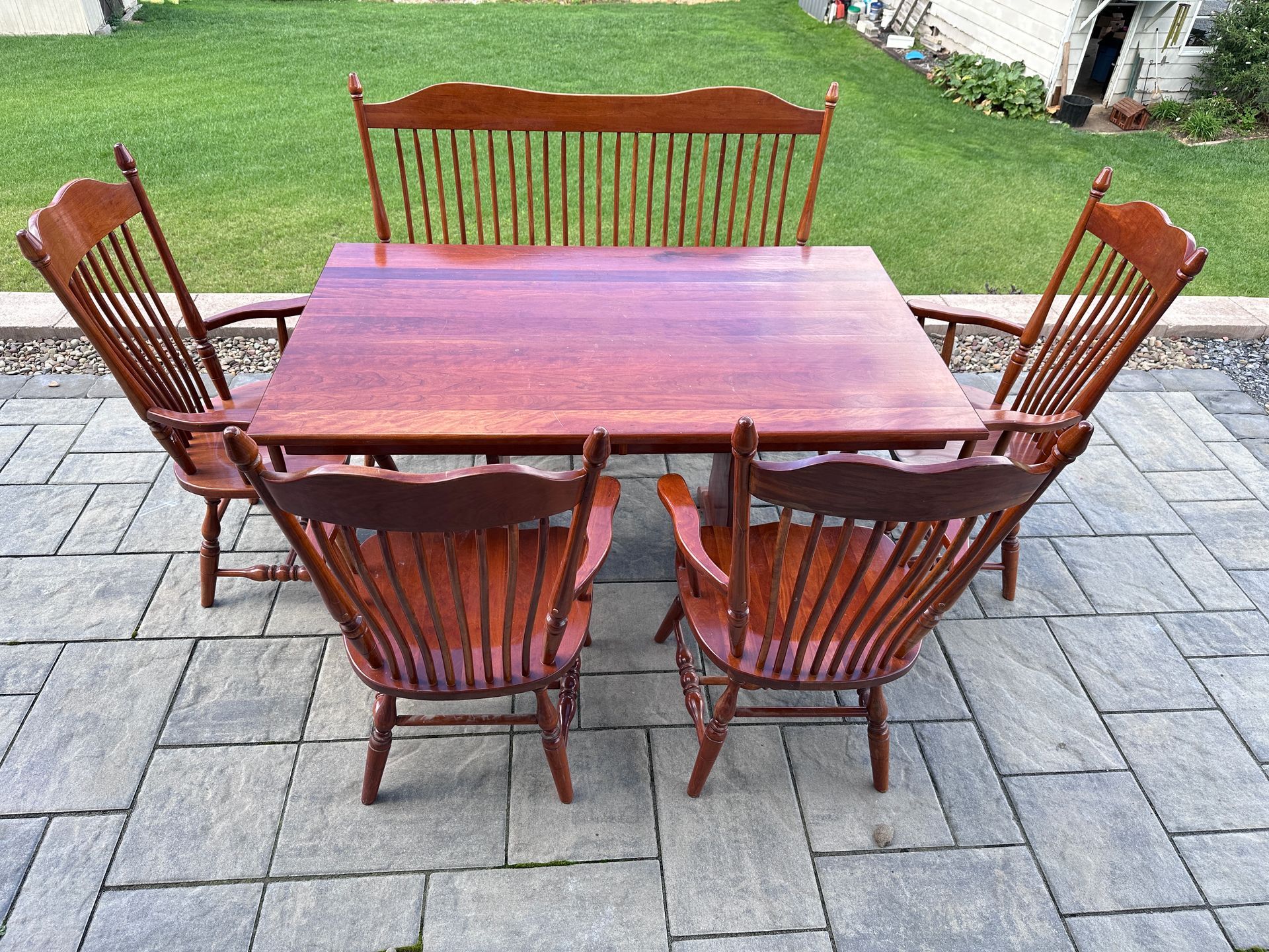 Wooden patio table surrounded by five chairs on a paved backyard patio with grass beyond.