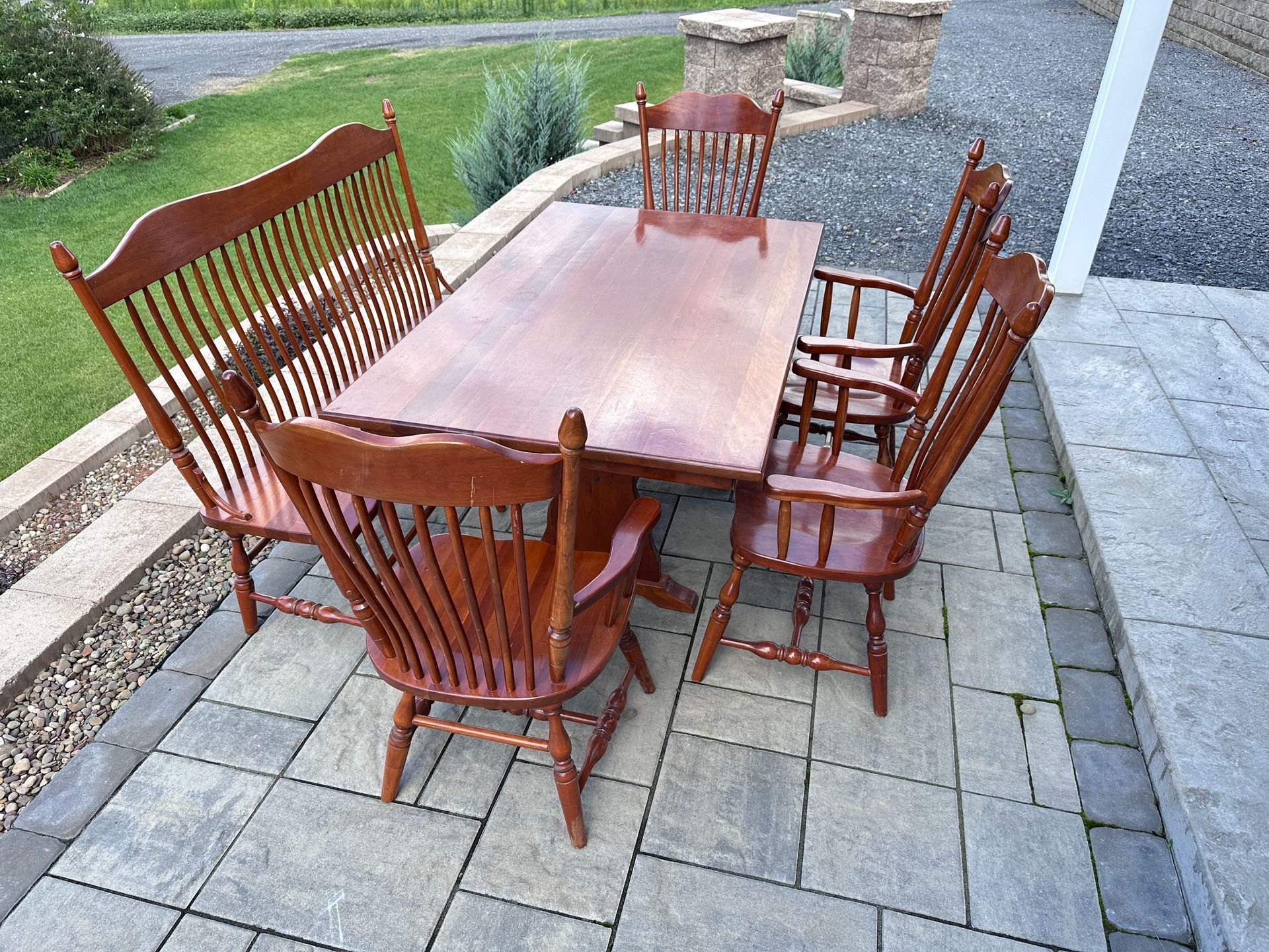 Wooden outdoor dining table with six chairs on a stone patio near a garden and gravel path