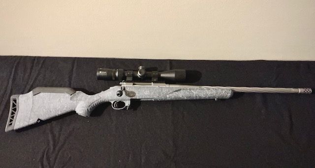A camouflage bolt-action rifle with scope, resting on a dark couch against a light wall.