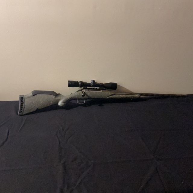Rifle with scope resting on a dark bedspread against a plain wall.