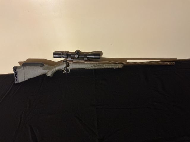 Scoped rifle on a black cloth backdrop with a beige wall behind it.