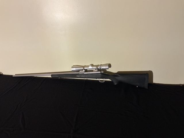 Rifle with scope resting on a black surface against a plain beige wall