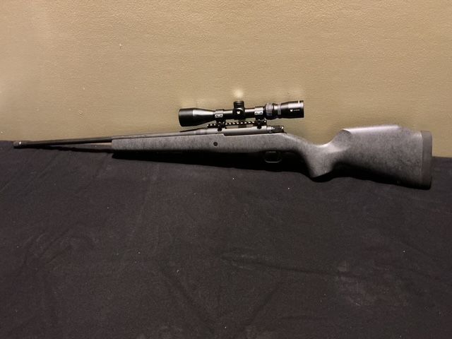 Black rifle with mounted scope on a dark fabric surface, viewed from the side.