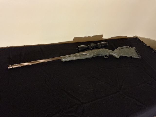 Rifle with scope and camouflage stock lying on a black cloth surface against a beige wall