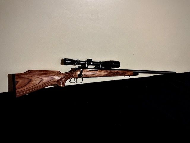 Bolt-action rifle with scope resting on a dark surface against a plain wall