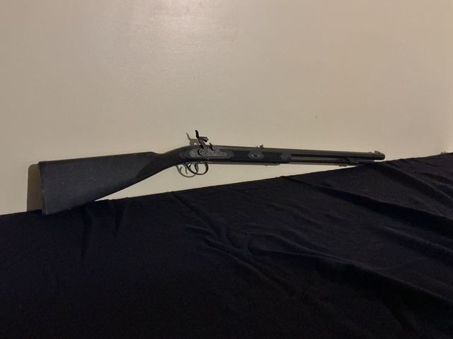 Black rifle resting on a draped black cloth, with a plain beige wall behind it