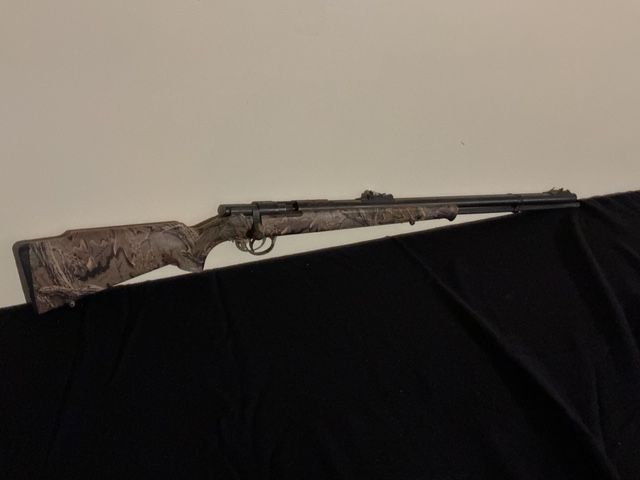 A camouflaged hunting rifle resting on a black draped surface against a plain wall.