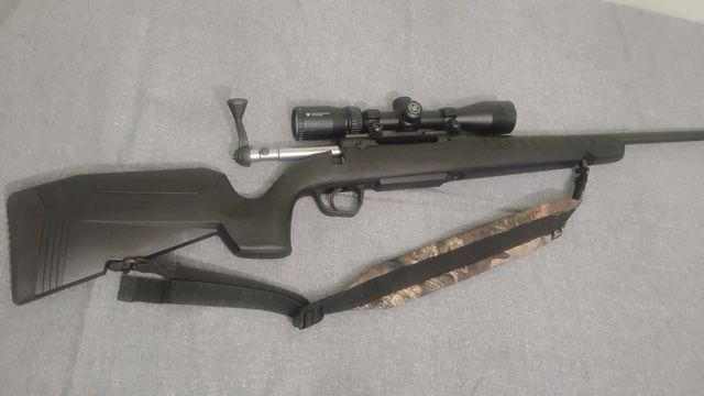 Bolt-action rifle with scope and black sling lying on a light gray surface