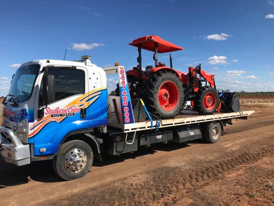 Blue flatbed truck carrying a red tractor on a dirt road under a clear sky