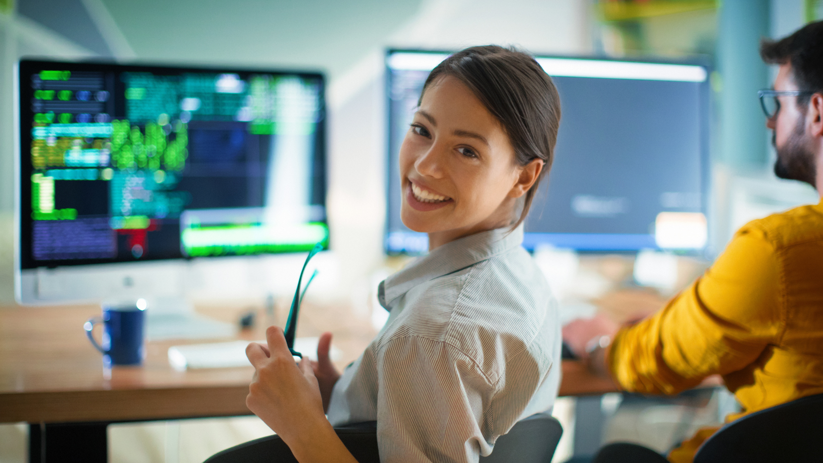 Woman smiling, holding glasses, working at a computer with a male colleague. Office setting, monitors display data.
