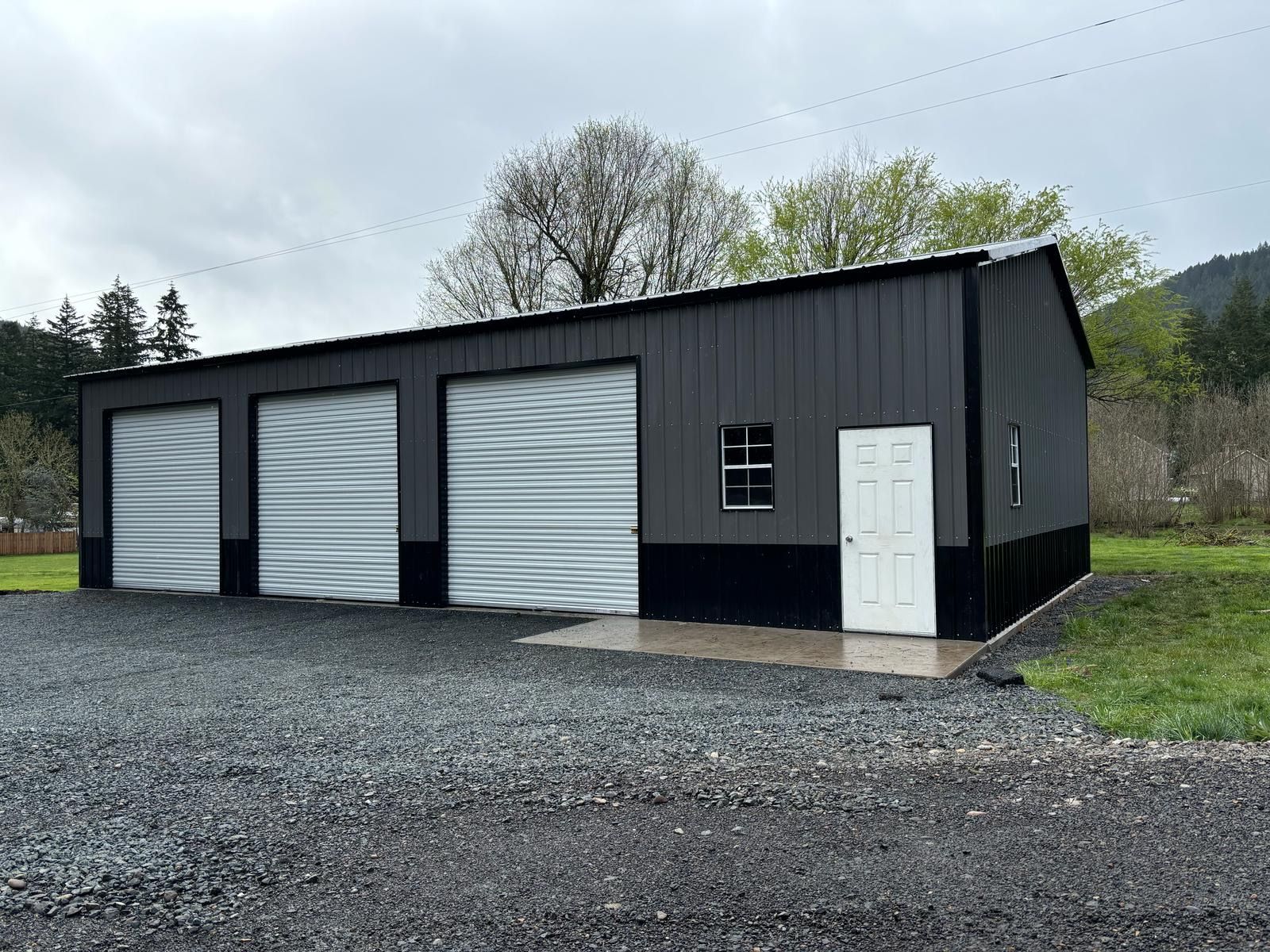 Black metal garage with three white roll-up doors and a side entrance on a gravel lot.