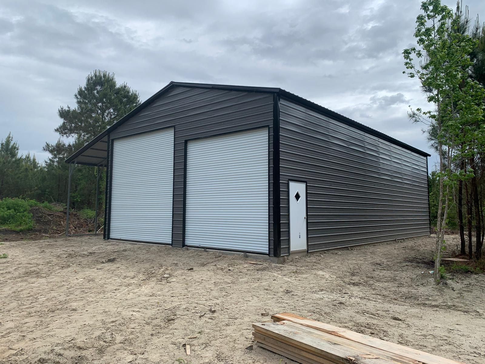 Dark gray metal garage with two white roll-up doors on a dirt lot under cloudy sky