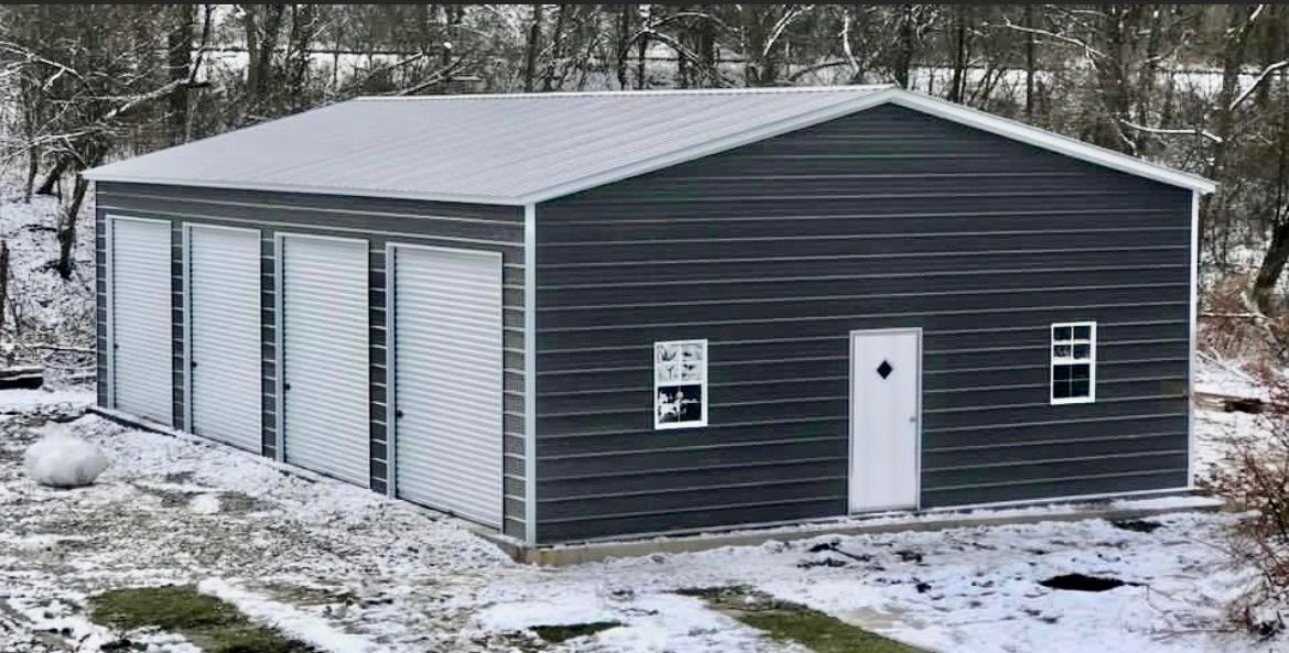 Dark gray metal garage building with white doors in a snowy wooded yard