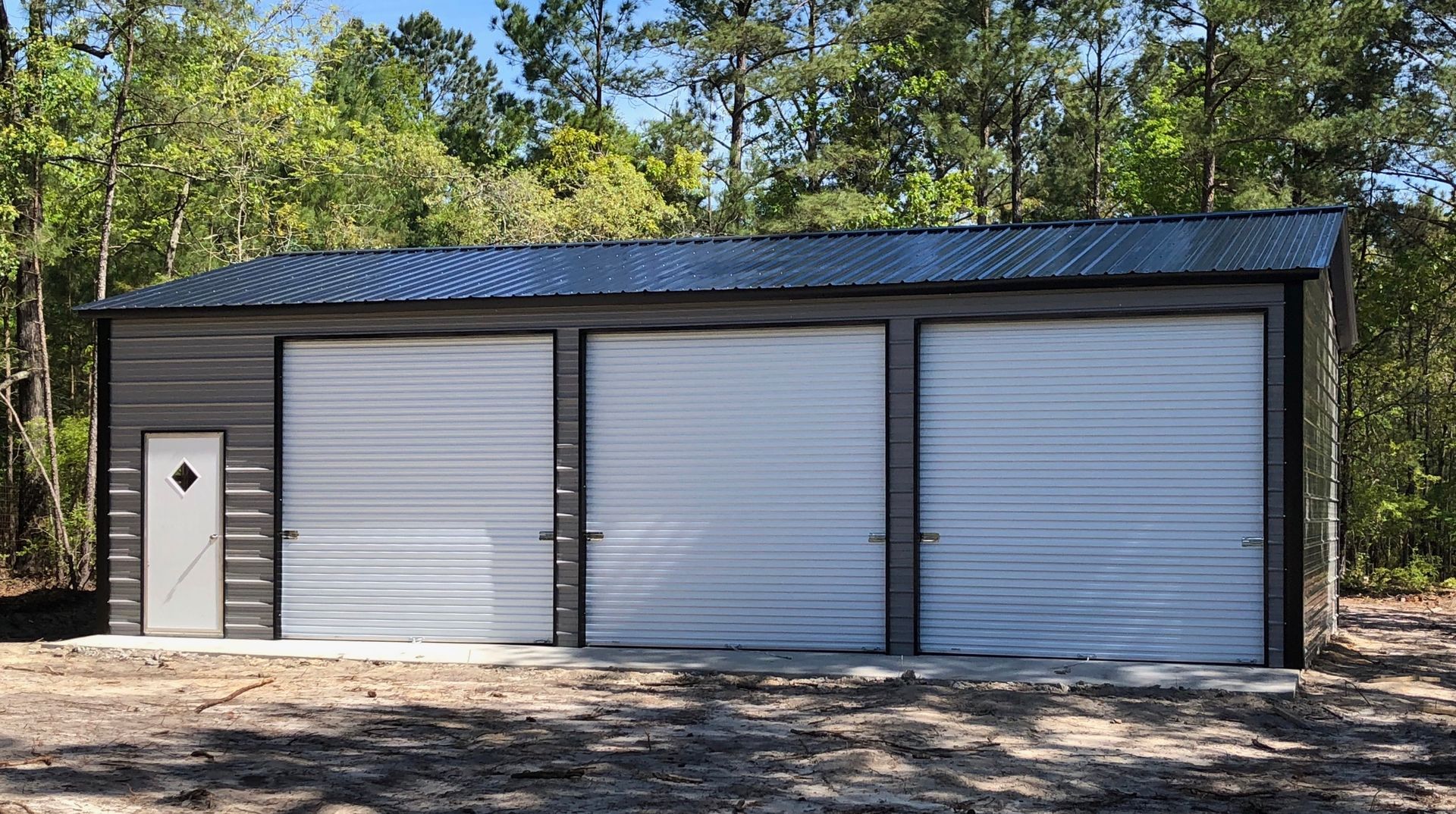 Three-bay metal garage with white doors and a side entry door in a wooded setting.