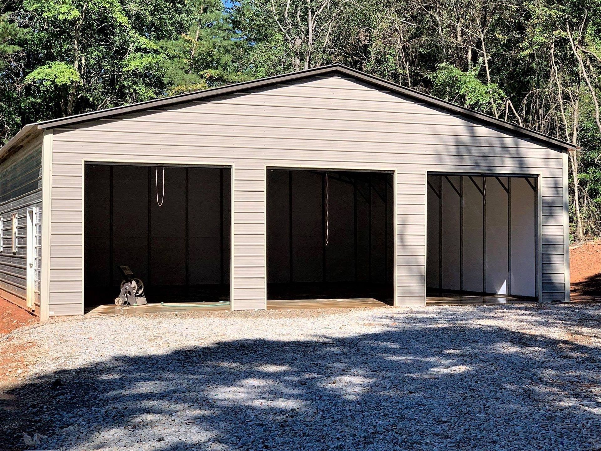 Three-bay tan garage with dark open doors and a gravel driveway, backed by trees.