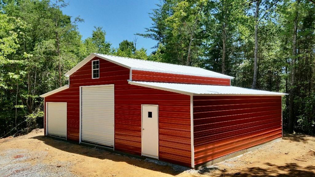 Red metal barn with white trim and large garage doors beside a dirt clearing and trees