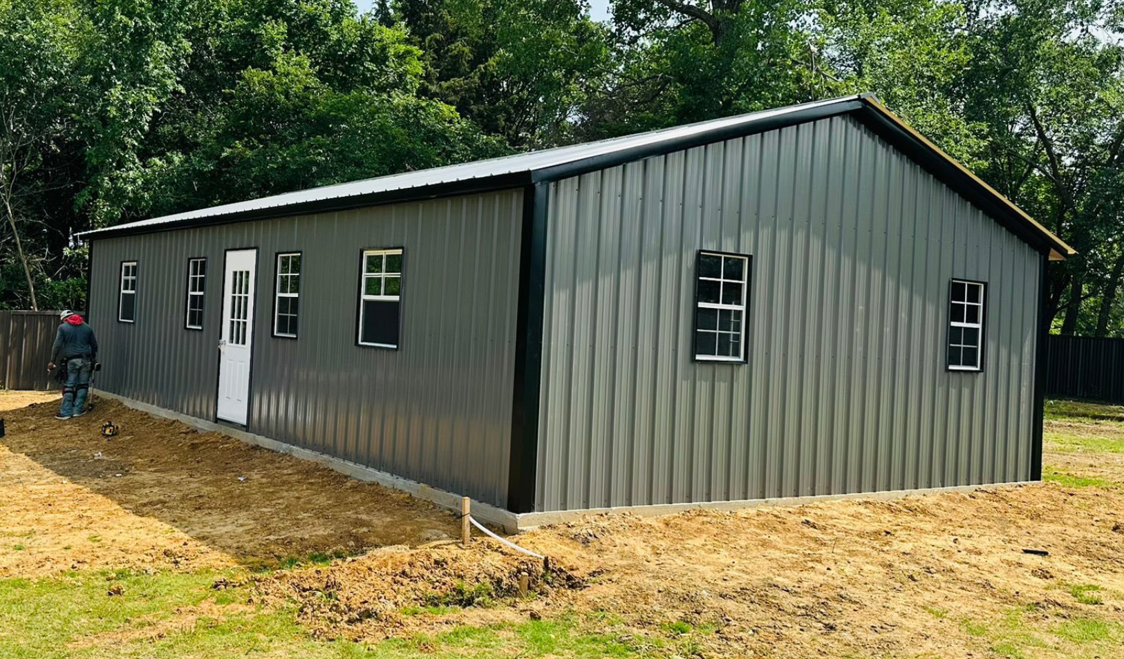 Gray metal building with multiple windows and a door, set on a dirt lot beside trees