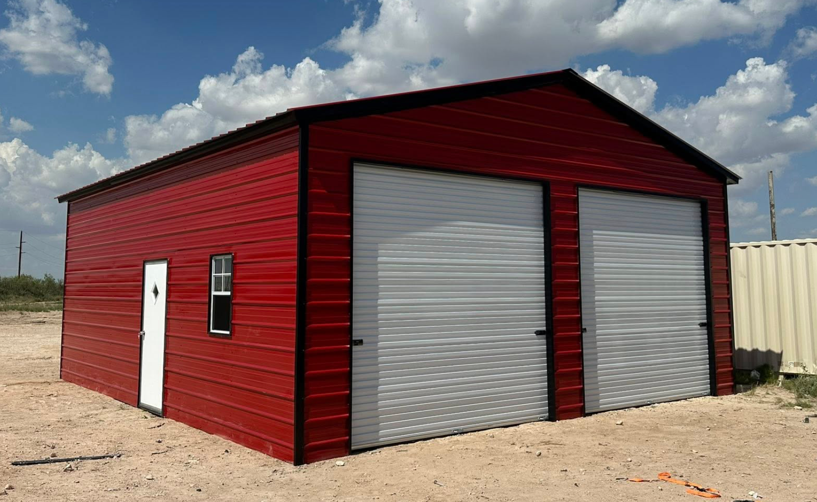 Red metal garage with two gray roll-up doors on a gravel lot under a cloudy sky