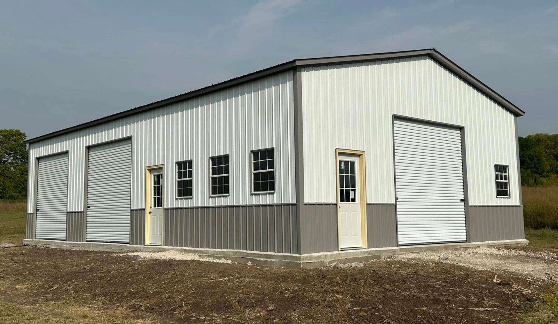 White metal warehouse building with roll-up doors and windows on a gravel lot under a cloudy sky